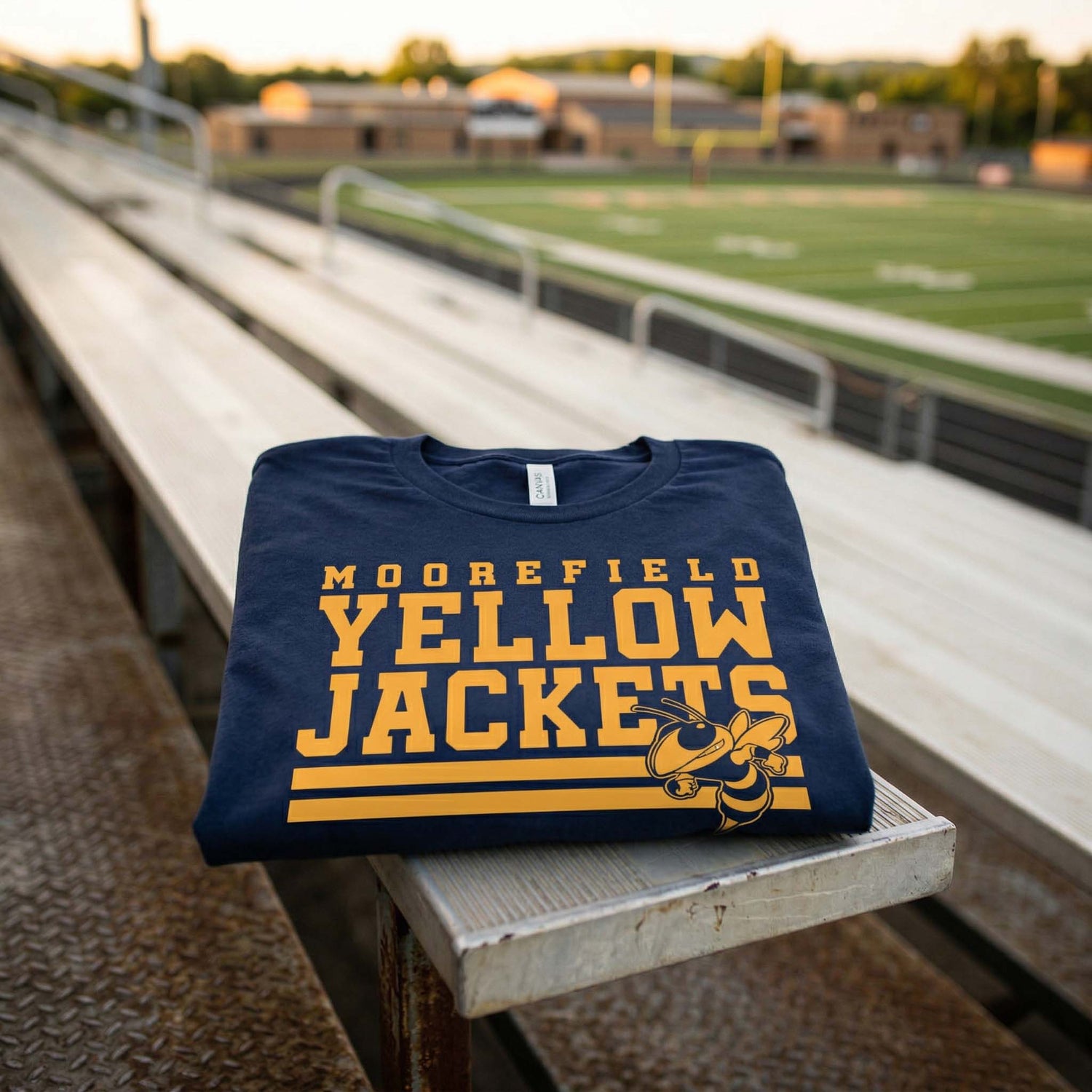 A navy blue Moorefield Yellow Jackets t-shirt with a bold gold graphic and hornet mascot, folded neatly on a wooden stadium bleacher with a blurred high school football field in the background at sunset.