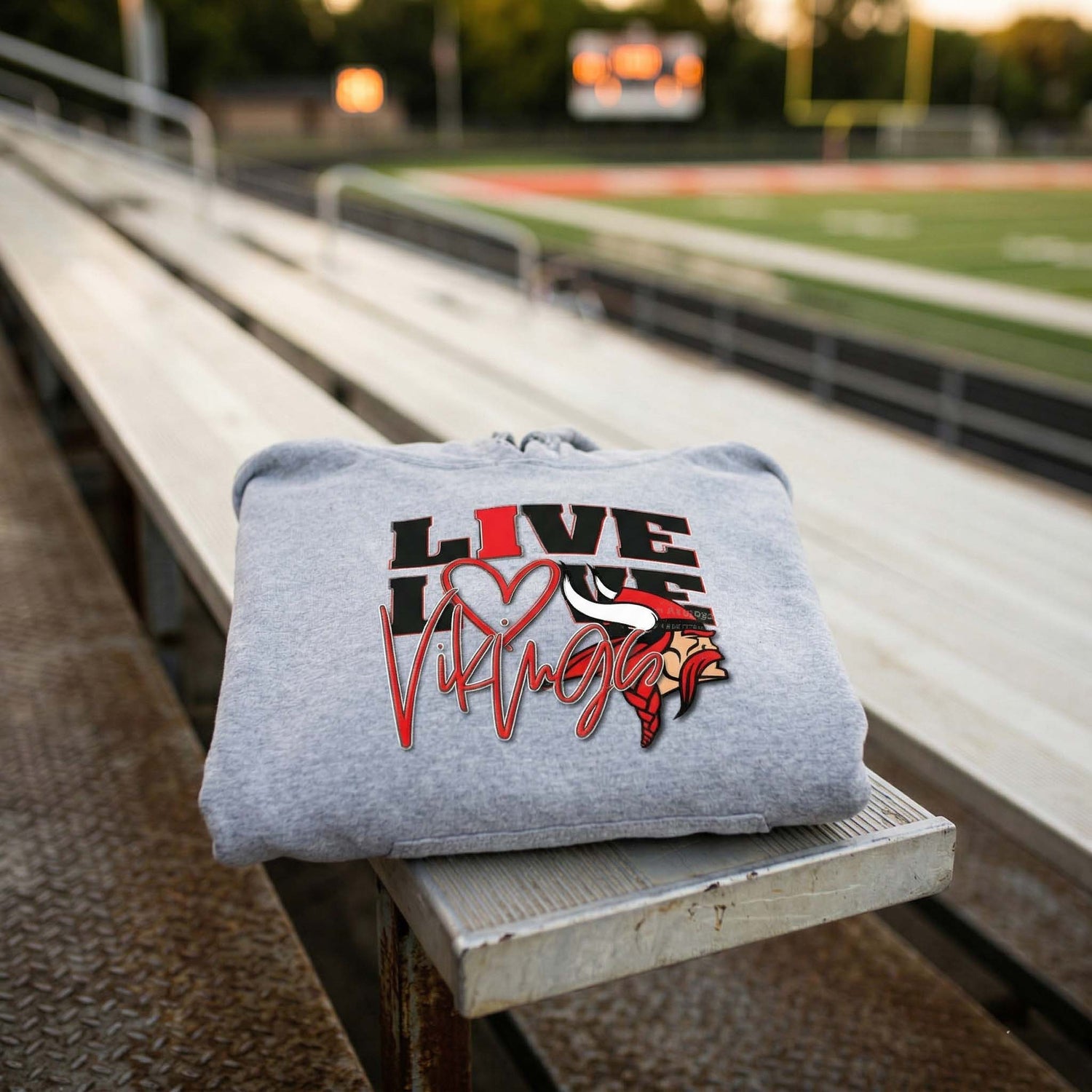 A heather grey Petersburg Vikings hoodie featuring a 'LIVE LOVE VIKINGS' graphic and red mascot, resting on wooden stadium bleachers with a blurred high school football field in the background.
