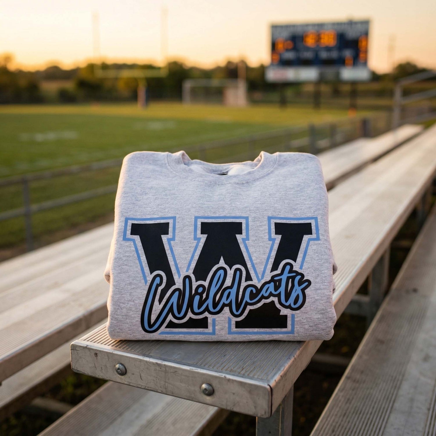A neatly folded light grey sweatshirt featuring a bold black 'W' and blue 'Wildcats' script, sitting on aluminum stadium bleachers with a blurred football field and scoreboard in the golden hour background.