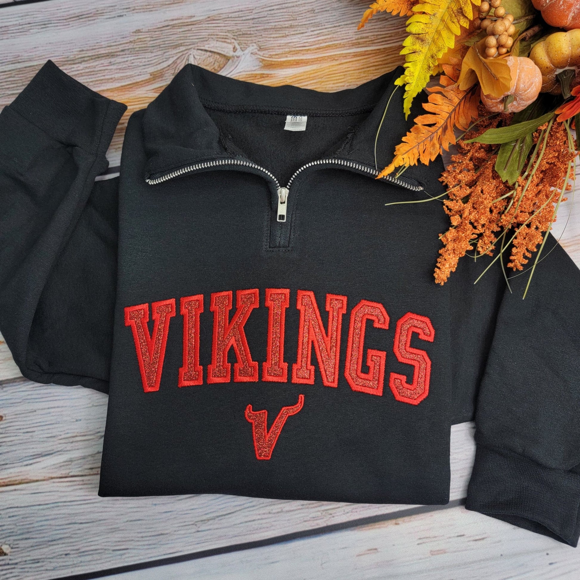 Black zip-up sweatshirt with 'VIKINGS' in red text on a wooden surface with autumn decorations.