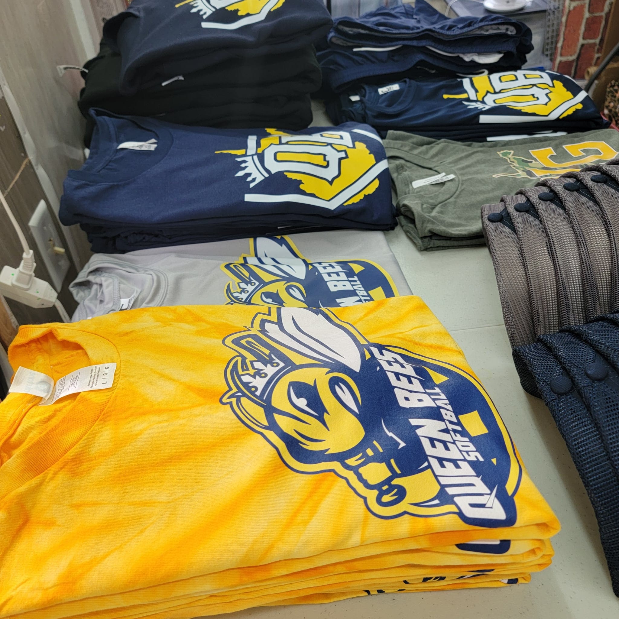 Stack of folded t-shirts with a prominent Queen bees logo on a table.