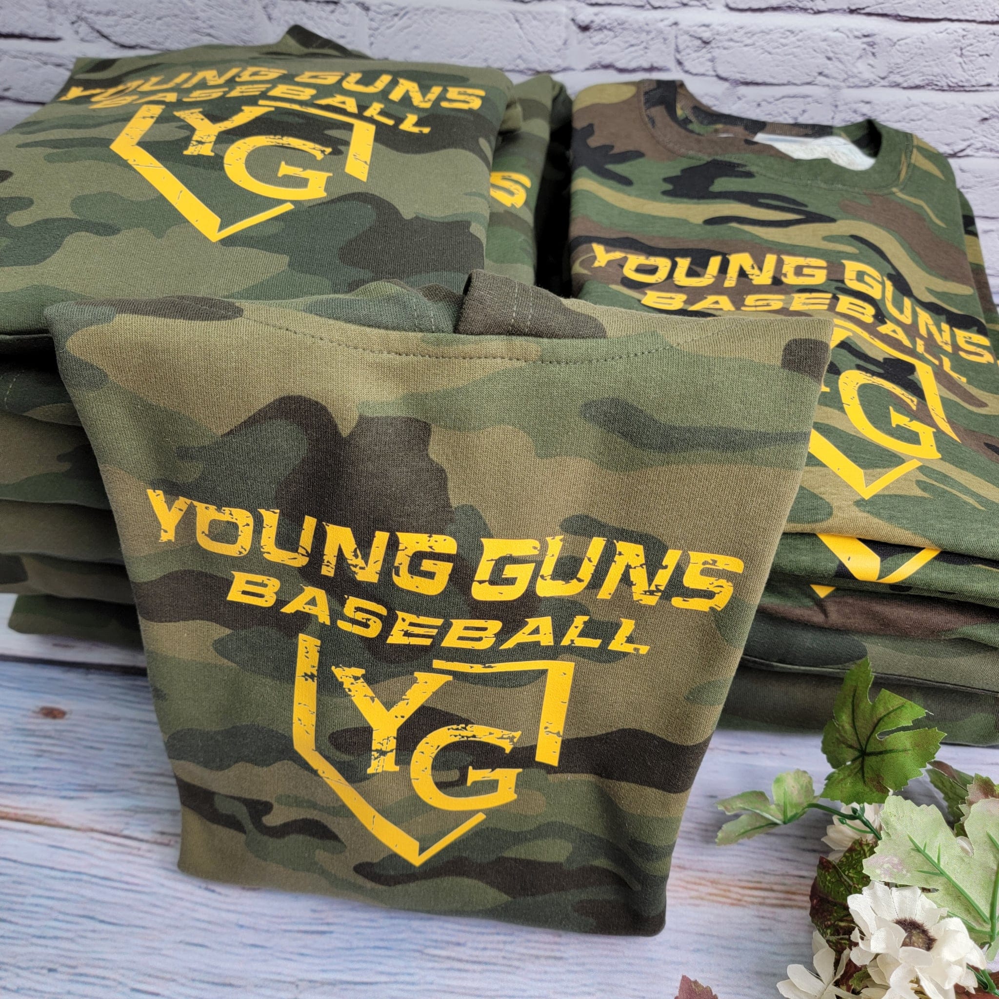 Stack of camouflage t-shirts with 'Young Guns Baseball' text on a wooden surface.